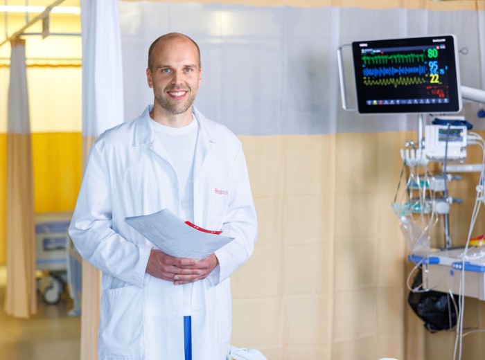 Dr Martin Serg soovitab elustiili tervislikumaks muutmisega alustada juba täna ning seada endale selleks teostatavad eesmärgid. Foto: erakogu meesterahvas seisab valges kitlis, käes on paberid, taustal palati kardinad ja ekraanid.